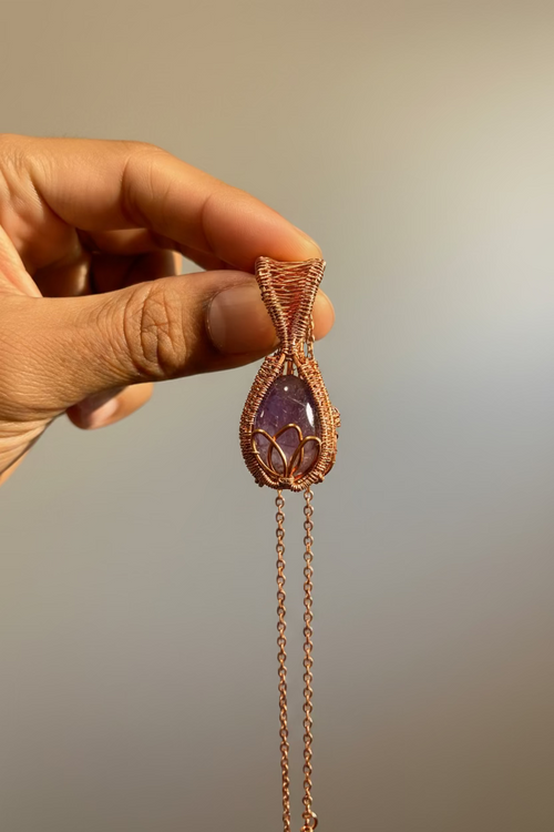 Hand holding a copper-wrapped amethyst pendant against a neutral background