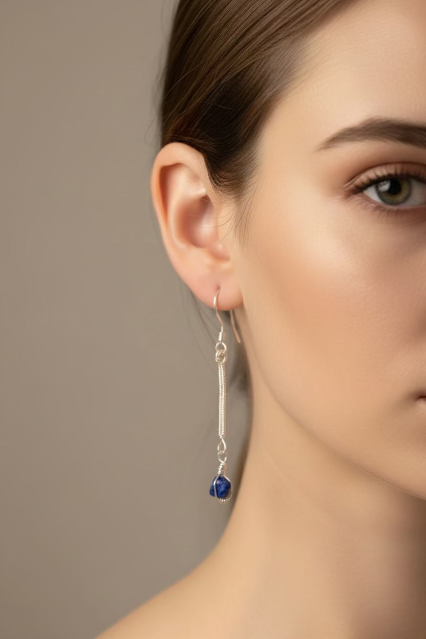 A close-up of a woman's ear and side profile wearing a long silver dangle earring with a small dark blue bead at the end, against a soft beige background.