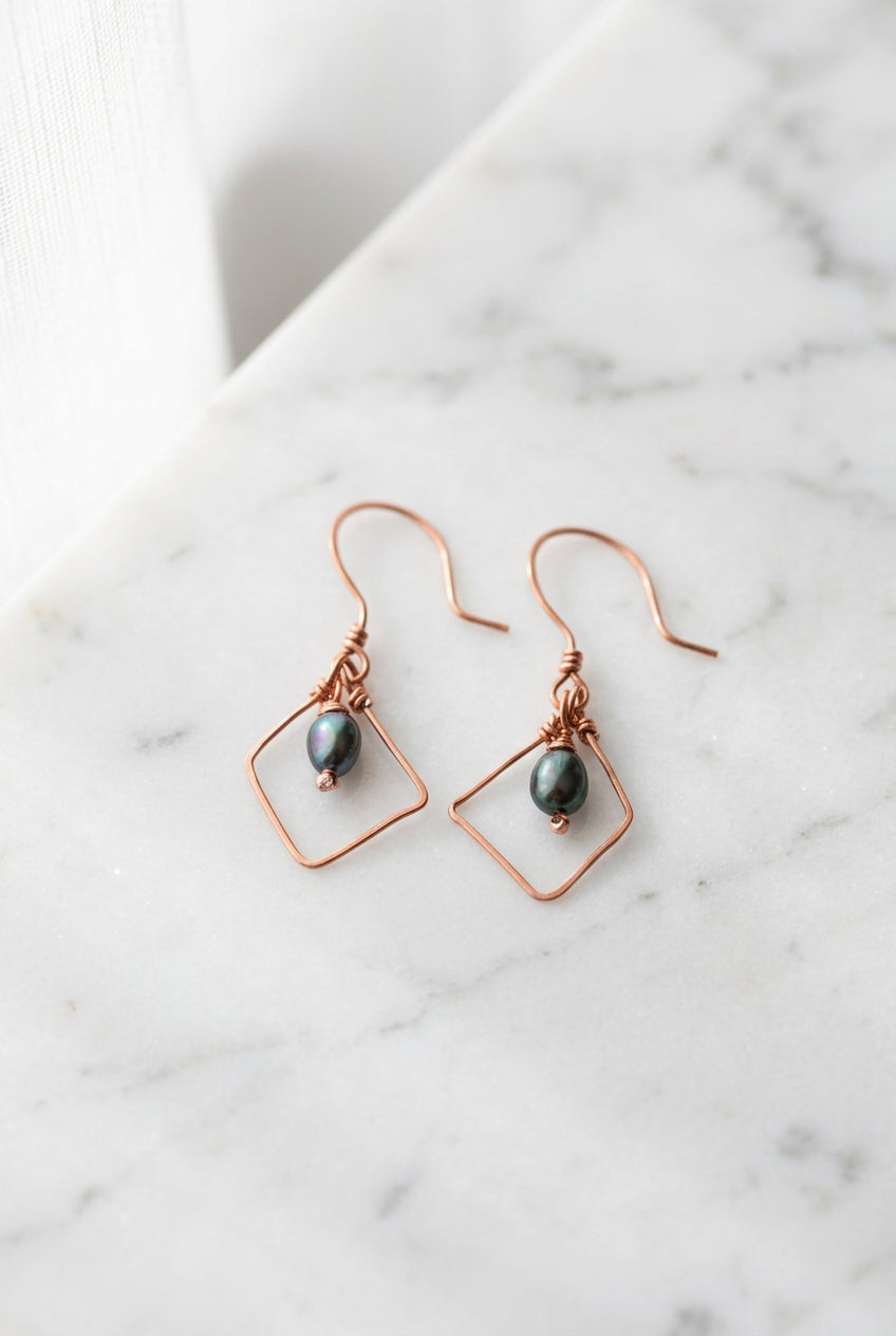 Pearl Alchemy geometric dangle earrings featuring dark blue/grey pearls suspended within diamond-shaped copper wire frames.