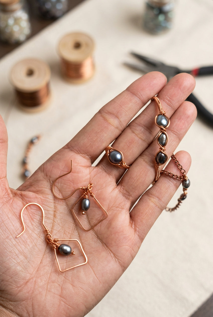 Hand holding Pearl Alchemy jewelry featuring a copper wire-wrapped grey pearl bracelet, matching pearl ring, and geometric copper dangle earrings, with copper wire spools and jewelry tools in the background.