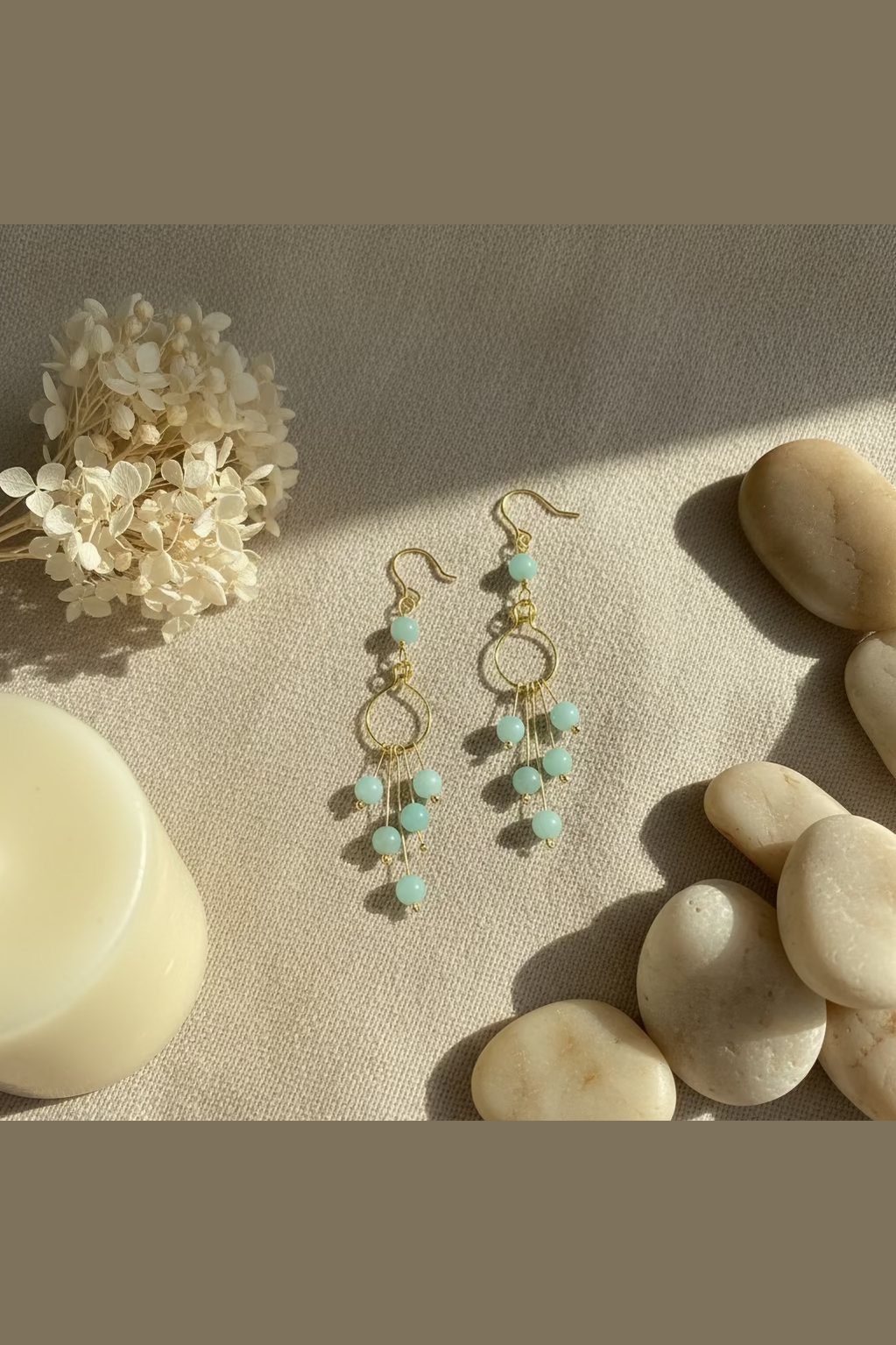 Flat lay of Seafoam Serenade handmade Amazonite earrings in golden-toned copper styled with smooth stones and dried hydrangea in warm natural sunlight.